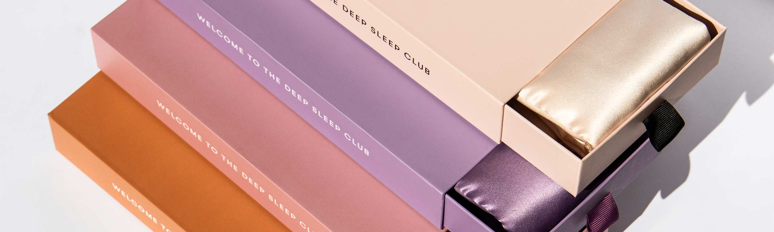 Four rectangular boxes in pastel peach, purple, and rust colors, each labeled “WELCOME TO THE DEEP SLEEP CLUB”, contain shiny satin sleep masks in matching shades, partially visible from open drawers.