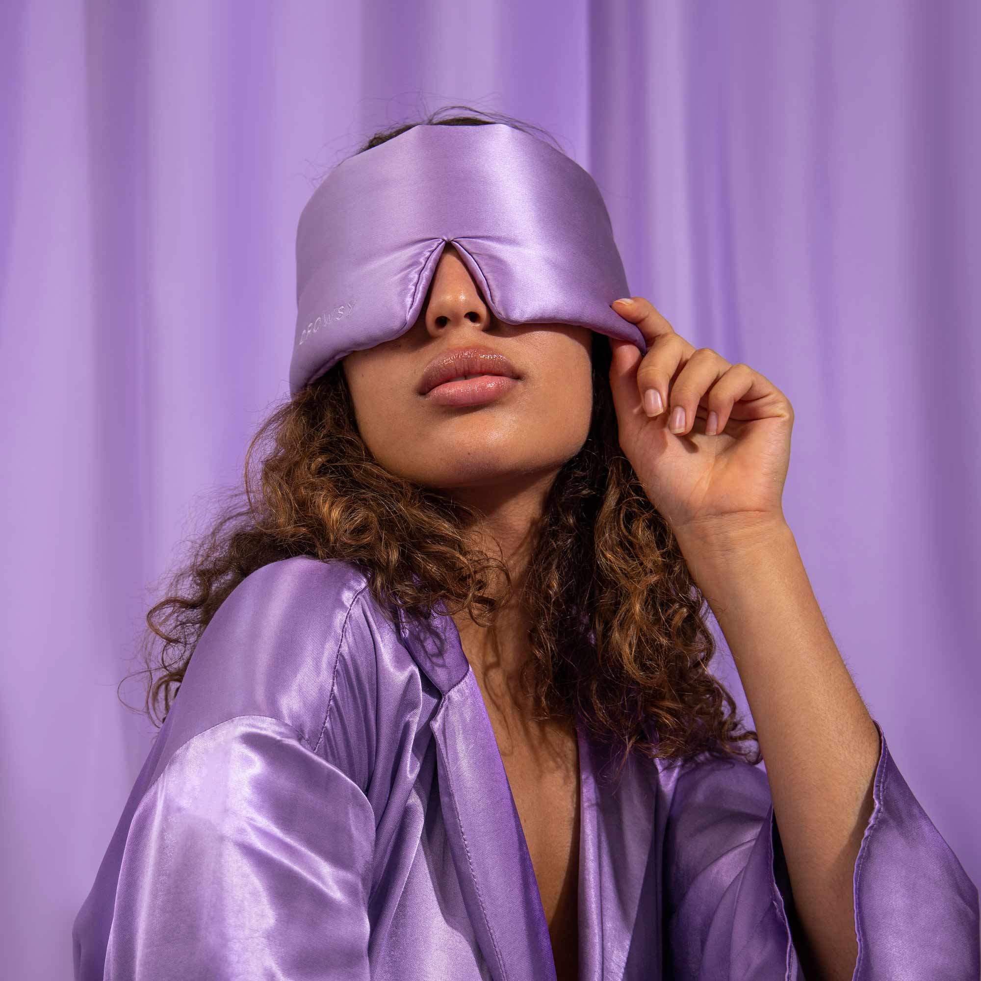 A model showcases the drowsysleepco Bundle - Lavender Haze, wearing the shiny lavender robe and eye mask against a matching background, elegantly lifting the mask with one hand for a touch of mystery.