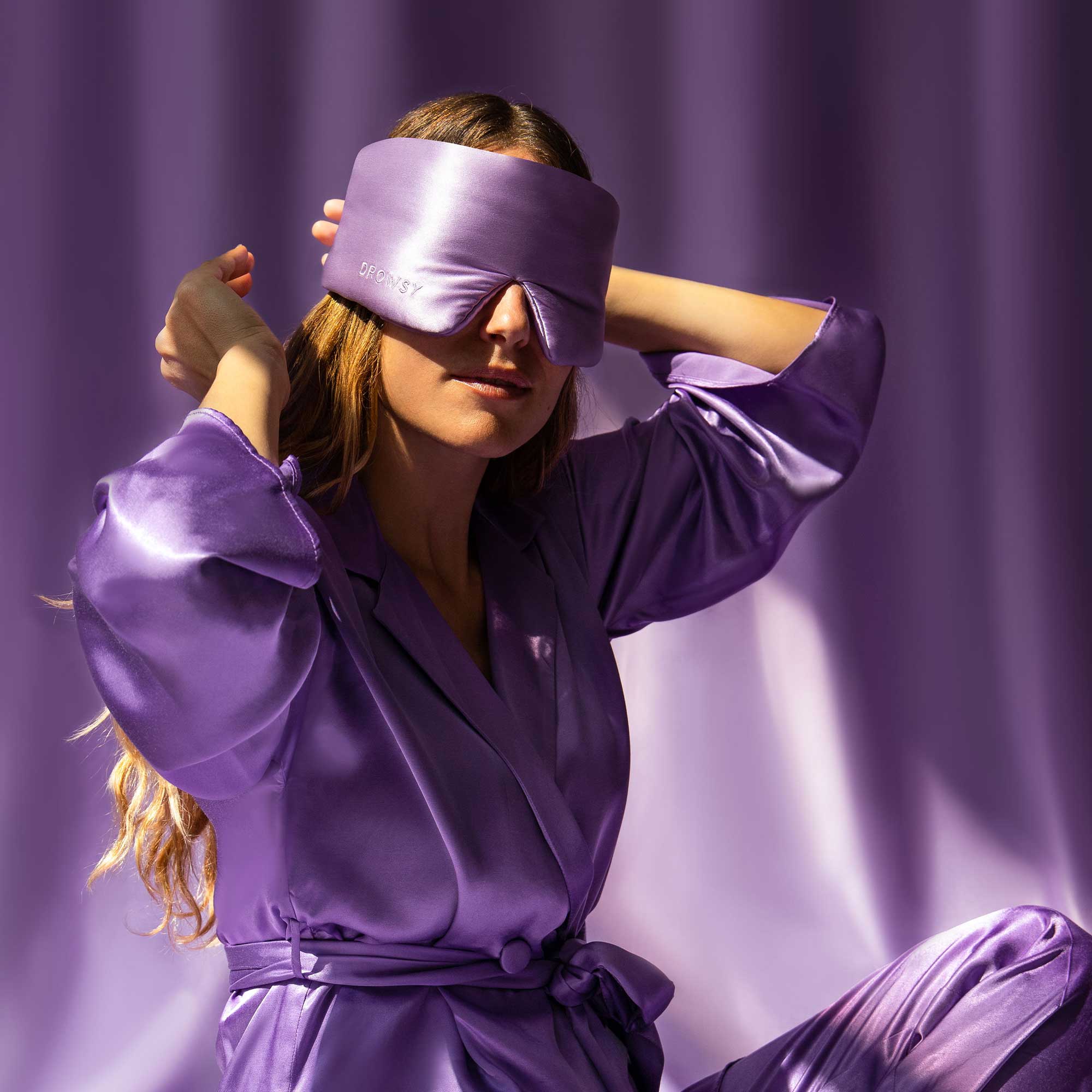 A woman in drowsysleepcos Lavender Haze satin robe and sleep mask poses before a purple backdrop, adjusting the mask as soft light highlights the silky fabric—perfect for ultimate beauty sleep.