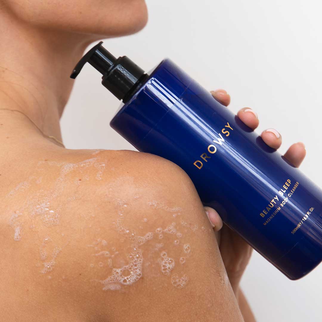 A person holds a large blue bottle labeled Beauty Sleep Ritual by drowsysleepco against their soapy shoulder, suggesting use of the magnesium body care product while bathing.