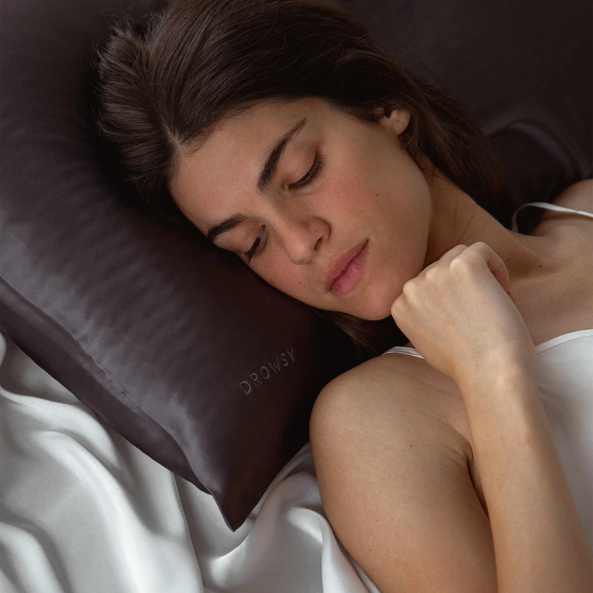 A woman with long brown hair sleeps peacefully on a dark-colored pillow labeled DROWSY, resting on white satin sheets. She is wearing a white sleeveless top.