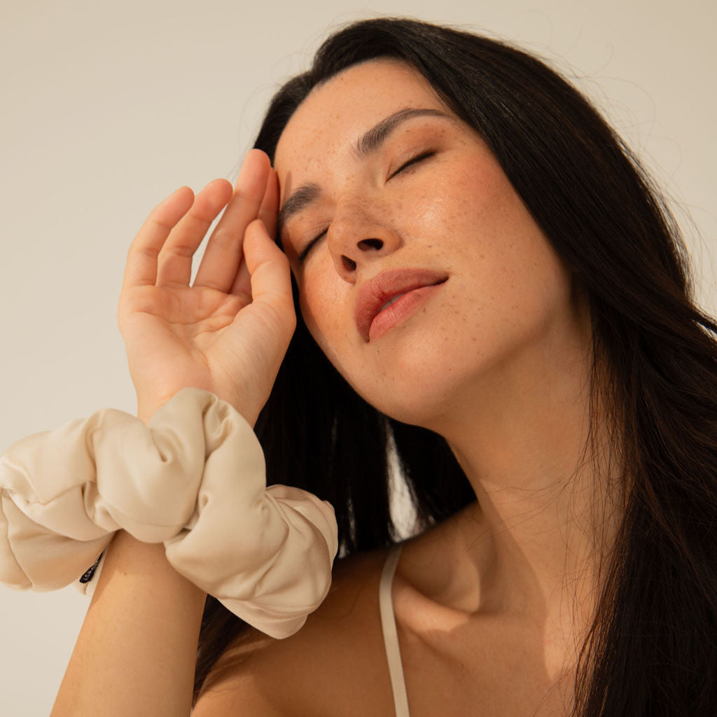 A woman with long dark hair and freckles closes her eyes and gently touches her forehead with her hand. She wears a cream-colored scrunchie on her wrist and a light camisole, appearing relaxed against a neutral background.