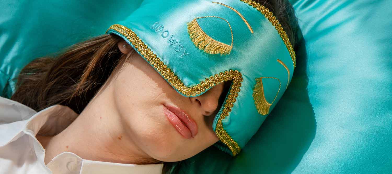 A woman reclines wearing the drowsysleepco Breakfast at Drowsy collection (original) bundle: a turquoise silk sleep mask with gold trim, matching satin pillow, and coordinating silk scrunchie.