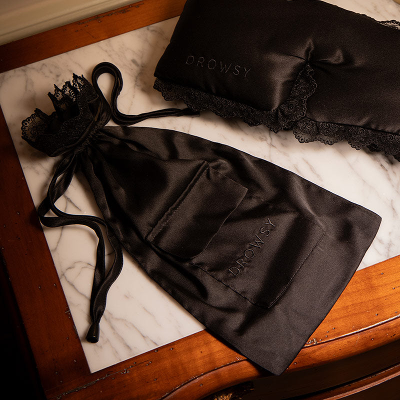 A black silk sleep mask with couture-inspired lace trim and a matching Paris Love Story Carry Pouch from drowsysleepco rest on a marble-topped wooden nightstand, evoking the elegance of a Parisian romance.