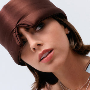 A woman with short brown hair wears the Rajah Sleep Mask by drowsysleepco, a silky brown eye mask that covers part of her forehead and one eyebrow as she gazes slightly upward with a neutral expression and glossy lips.