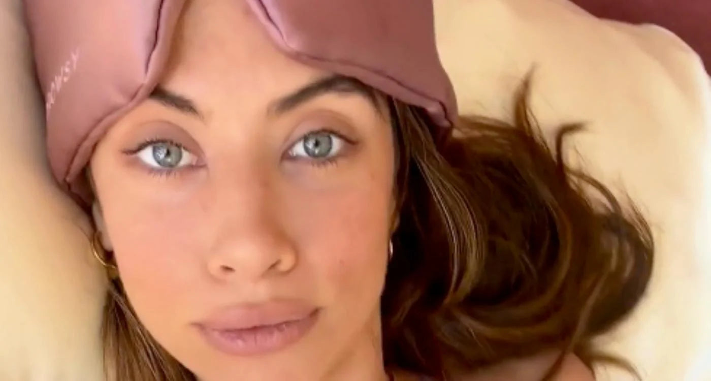 A woman with light eyes and long brown hair lies on a pillow, wearing a mauve sleep mask pushed up on her forehead. She looks directly at the camera with a relaxed expression.