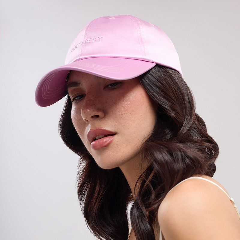 A woman with long, wavy dark hair wears the Drowsy Silk Cap - Hotel Paradiso Heatwave by Drowsy and a sleeveless top. She gazes slightly to the side with a neutral expression against a plain background.