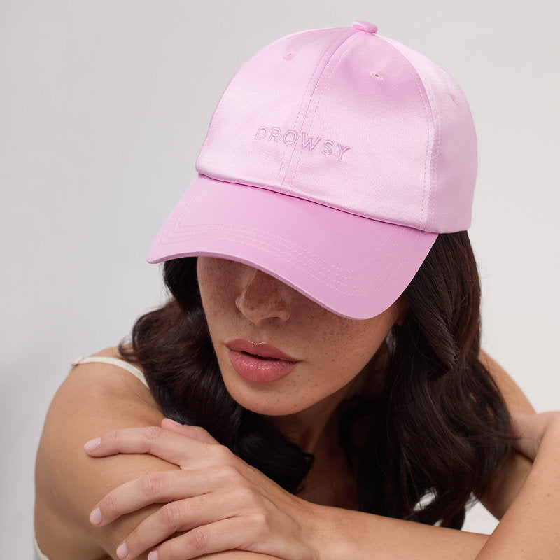 A woman with wavy brown hair wears the Drowsy Silk Cap - Hotel Paradiso Heatwave by Drowsy, featuring light pink silk and embroidered branding for stylish hair protection. She looks down with arms crossed and lips slightly parted.