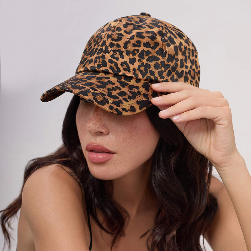 A woman with long dark hair wears the Drowsy Silk Cap - Hotel Paradiso Wild Thing by Drowsy, paired with a black top, tilting the cap's brim down with her right hand to partially shade her face from the sun.