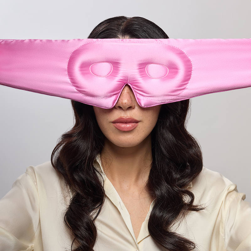 A woman with long dark hair, wearing a cream blouse, models the us.drowsysleepco Hotel Paradiso Heatwave Eyelash Protecting Mask, featuring eye holes. She faces the camera against a plain light background.
