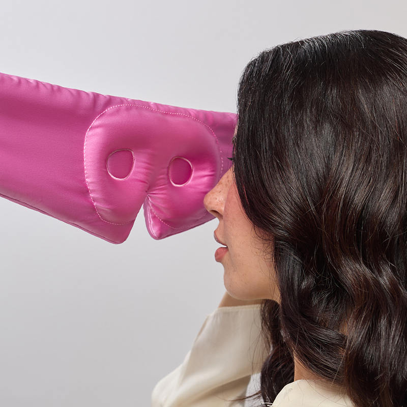 A woman with wavy dark hair holds the us.drowsysleepco Hotel Paradiso Heatwave Eyelash Protecting Mask, a pink mulberry silk sleep mask with cut-out eye holes, near her face as she prepares to put it on.
