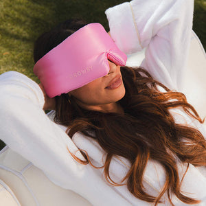 A woman in a white robe relaxes outside on a white chair, wearing the us.drowsysleepco Hotel Paradiso Heatwave Eyelash Protecting Mask, labeled DROWSY, enjoying total blackout comfort with her arms behind her head.