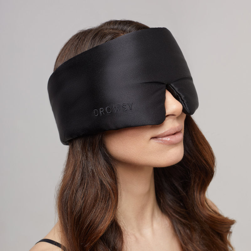 A woman with long brown hair wears the us.drowsysleepco Black Jade - Eyelash Protecting Mask, which covers her eyes and upper nose. She smiles slightly against a plain, light gray background.