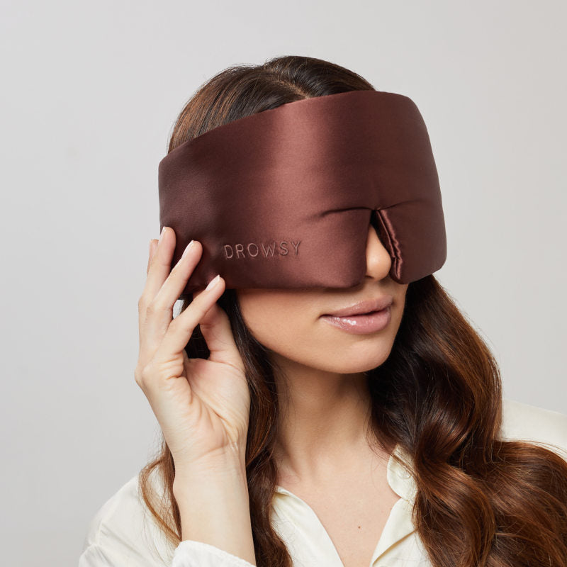 A woman with long brown hair gently touches the Cocoa Glow - Eyelash Protecting Mask by us.drowsysleepco, a large silky brown sleep mask. She wears a light-colored top.