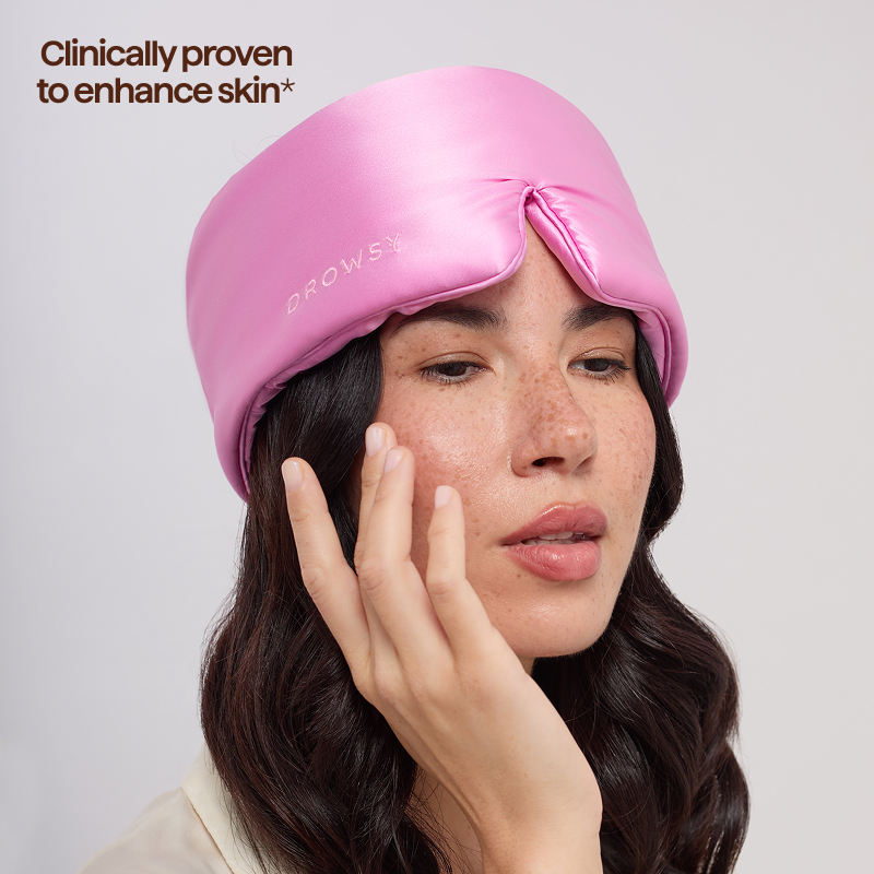 A woman with long dark hair wears the us.drowsysleepco Hotel Paradiso Heatwave Sleep Mask, gently touching her face. Text in the top left reads: Clinically proven to enhance skin*.