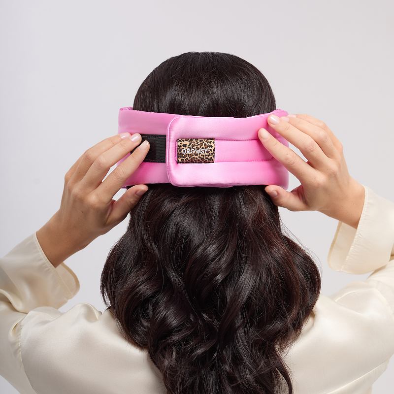A person with long, dark hair adjusts the us.drowsysleepco Hotel Paradiso Heatwave Eyelash Protecting Mask, featuring a pink mulberry silk band with a leopard print patch, against a plain light background.