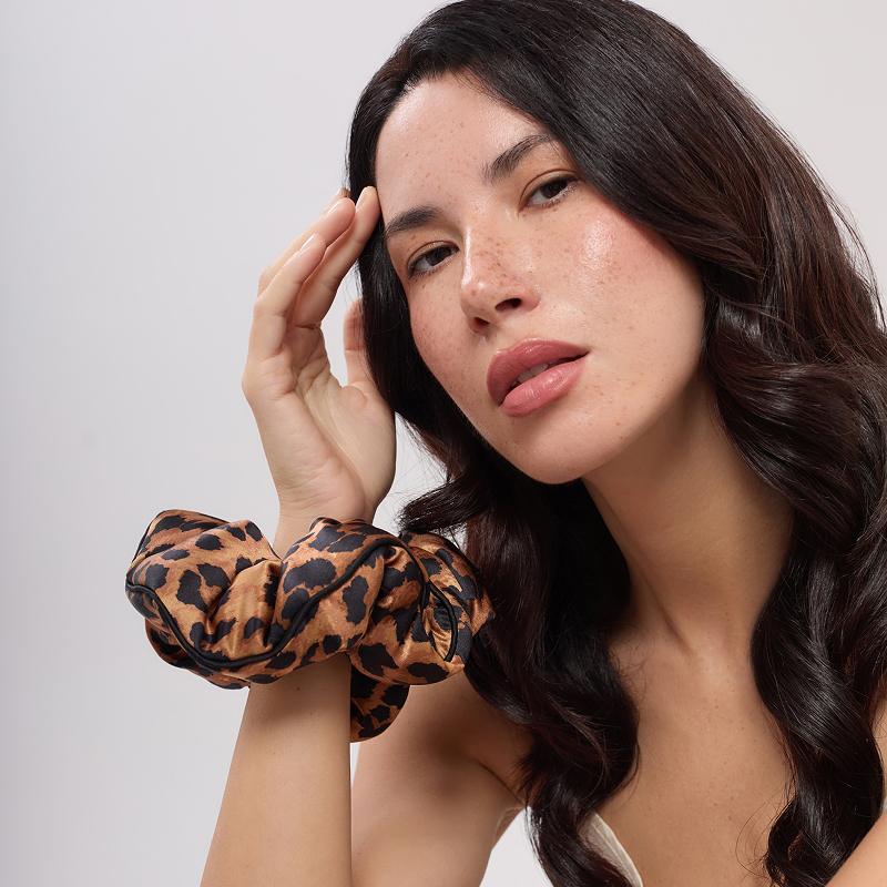 A woman with long, wavy dark hair poses confidently, wearing the Hotel Paradiso - Wild Thing Padded Scrunchie by us.drowsysleepco on her wrist. Styled with a white top against a plain background—showing off thickness and flair.