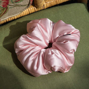 The us.drowsysleepco Petal Pink Padded Scrunchie, a large glossy pink pillow scrunchie, sits on green fabric by a wicker chair with a floral cushion—stylish and perfect for healthy hair.