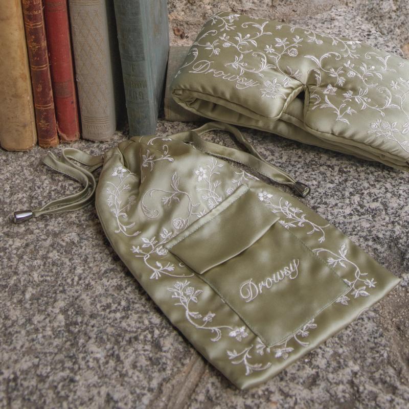 Pride & Prejudice - Sage Carry Pouch with a matching sleep mask, both featuring white floral embroidery and the word Drowsy, resting on a stone surface beside antique books.