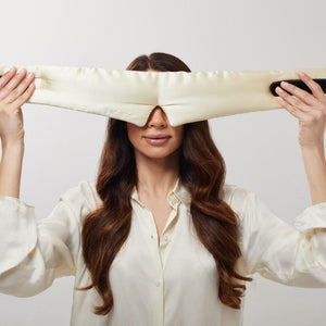 A woman with long brown hair in a white shirt holds the us.drowsysleepco Champagne Gold silk eye mask over her eyes, highlighting beauty sleep against a neutral background.