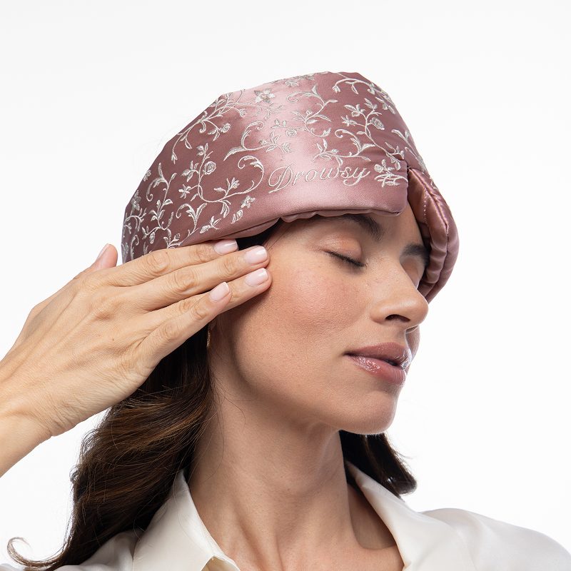 A person wearing a dusty pink Pride & Prejudice - English Rose Sleep Mask with silver floral embroidery over their eyes, with their hand touching the side of their face.