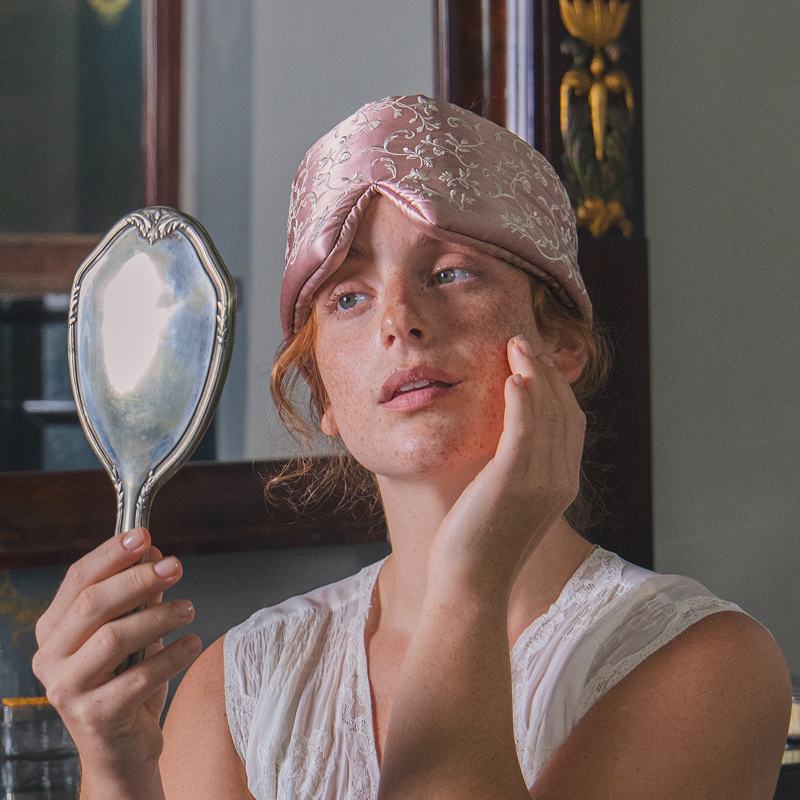 A person wearing a pale pink floral Pride & Prejudice - English Rose Sleep Mask on their forehead while looking into a handheld mirror.