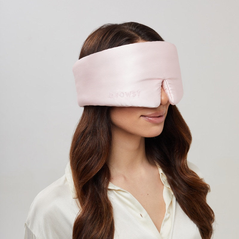 A woman with long brown hair wears the us.drowsysleepco Petal Pink adjustable sleep mask, which covers her eyes and nose. She is dressed in a white shirt and stands against a simple, light background.