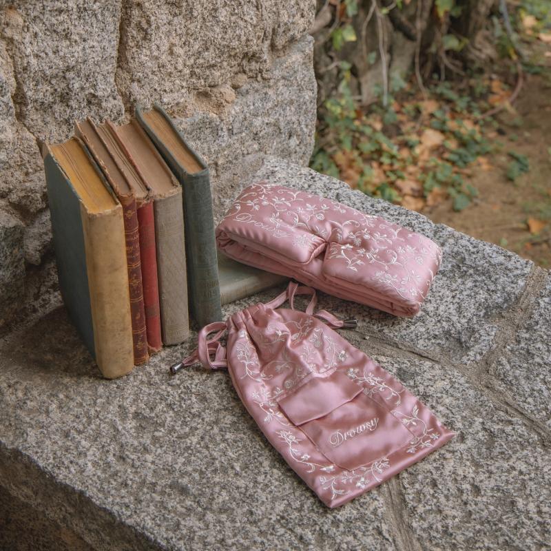 A pink Pride & Prejudice - English Rose Sleep Mask + Pouch made of mulberry silk with white floral embroidery, resting on a stone ledge next to a stack of vintage hardcover books.