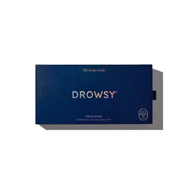 A minimalist rectangular dark blue box labeled “drowsysleepco” and “Midnight Blue - Swarovski® embellished logo” contains a silk sleep mask, featuring white and silver text about beauty sleep and a small circular emblem in the lower right corner.