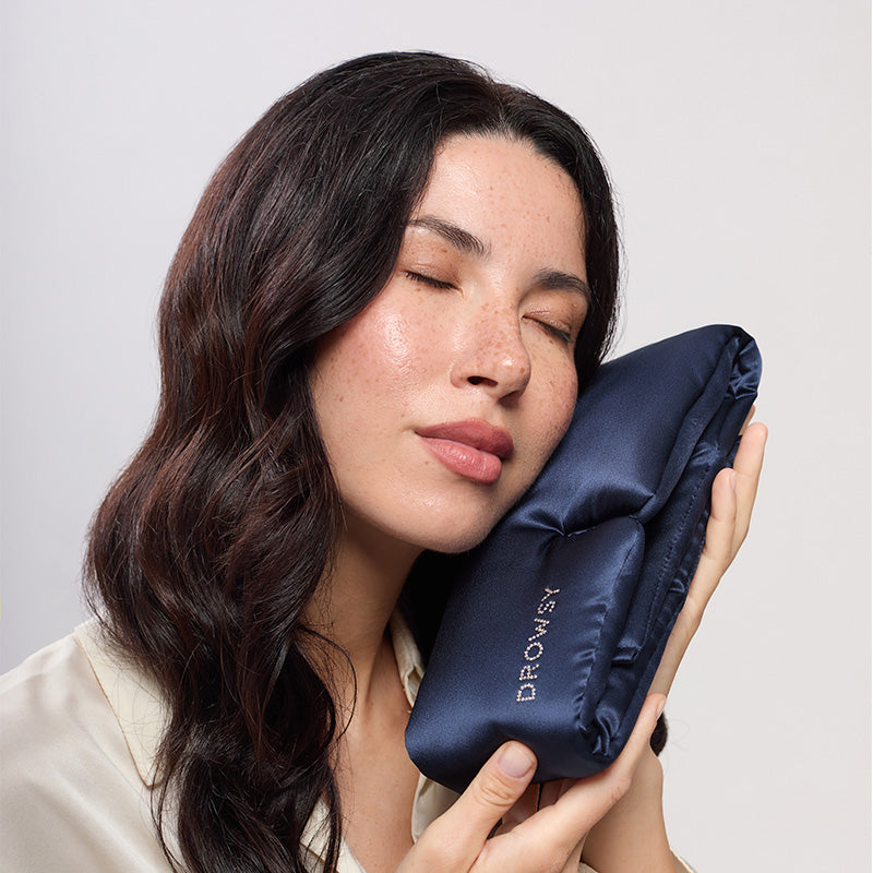 A woman with long dark hair smiles softly, eyes closed, as she rests her face on the drowsysleepco Midnight Blue silk sleep mask pillow featuring a Swarovski® embellished logo. She wears a light blouse against a plain background.