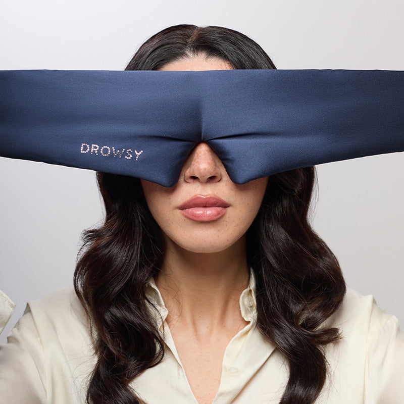 A woman with wavy dark hair wears the drowsysleepco Midnight Blue silk sleep mask with a Swarovski® embellished logo, covering her eyes and holding it at the sides. She is dressed in a light blouse against a plain background.