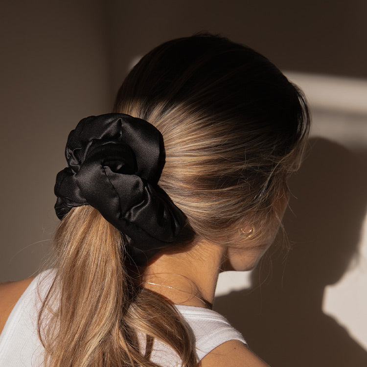 A woman with light brown hair in a low ponytail, secured by a large black satin scrunchie, stands in soft natural light. She pairs her white tank top with the drowsysleepco Sensual sleep bundle - Black Jade.