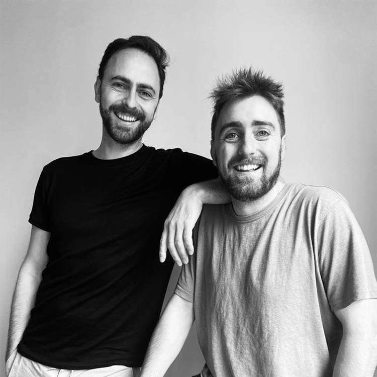Two men smiling at the camera, one wearing a black t-shirt and the other a light-colored t-shirt, standing close together against a plain background. The man in black has his arm resting on the other’s shoulder.