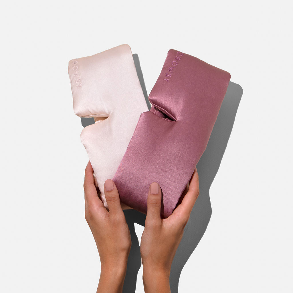 Two hands hold drowsysleepco’s Lovers Pack: two plush mulberry silk eye masks, one light pink and one mauve, set against a plain white background.