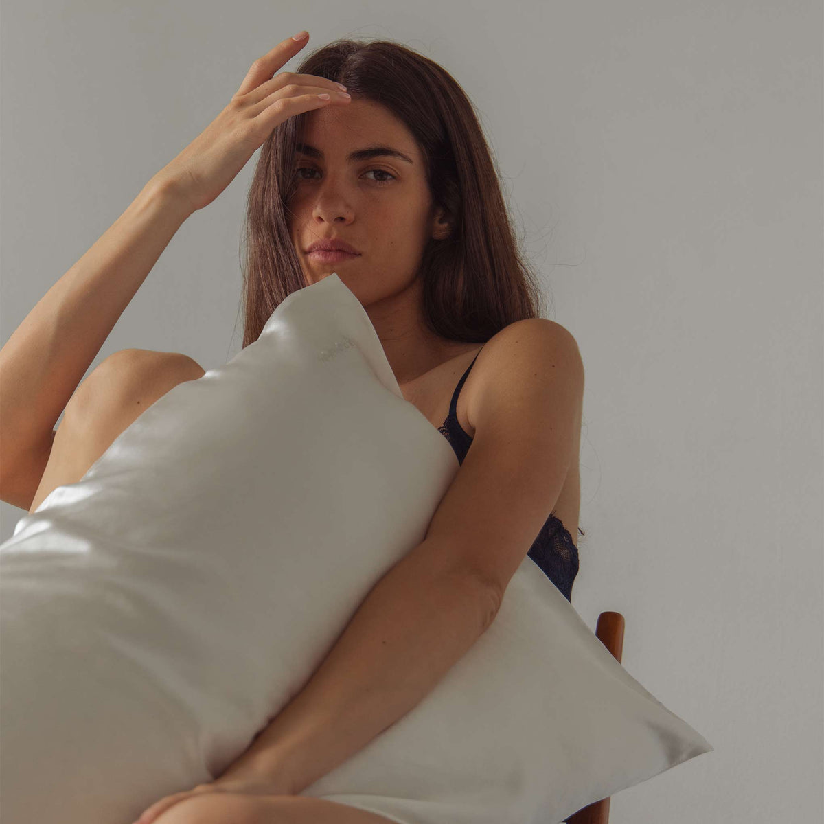 A woman with long brown hair sits on a chair, wearing a dark sleeveless top, holding the drowsysleepco Perfect Skin Bundle Pink Damask Rose pillowcase against her body and touching her forehead, against a plain light background.