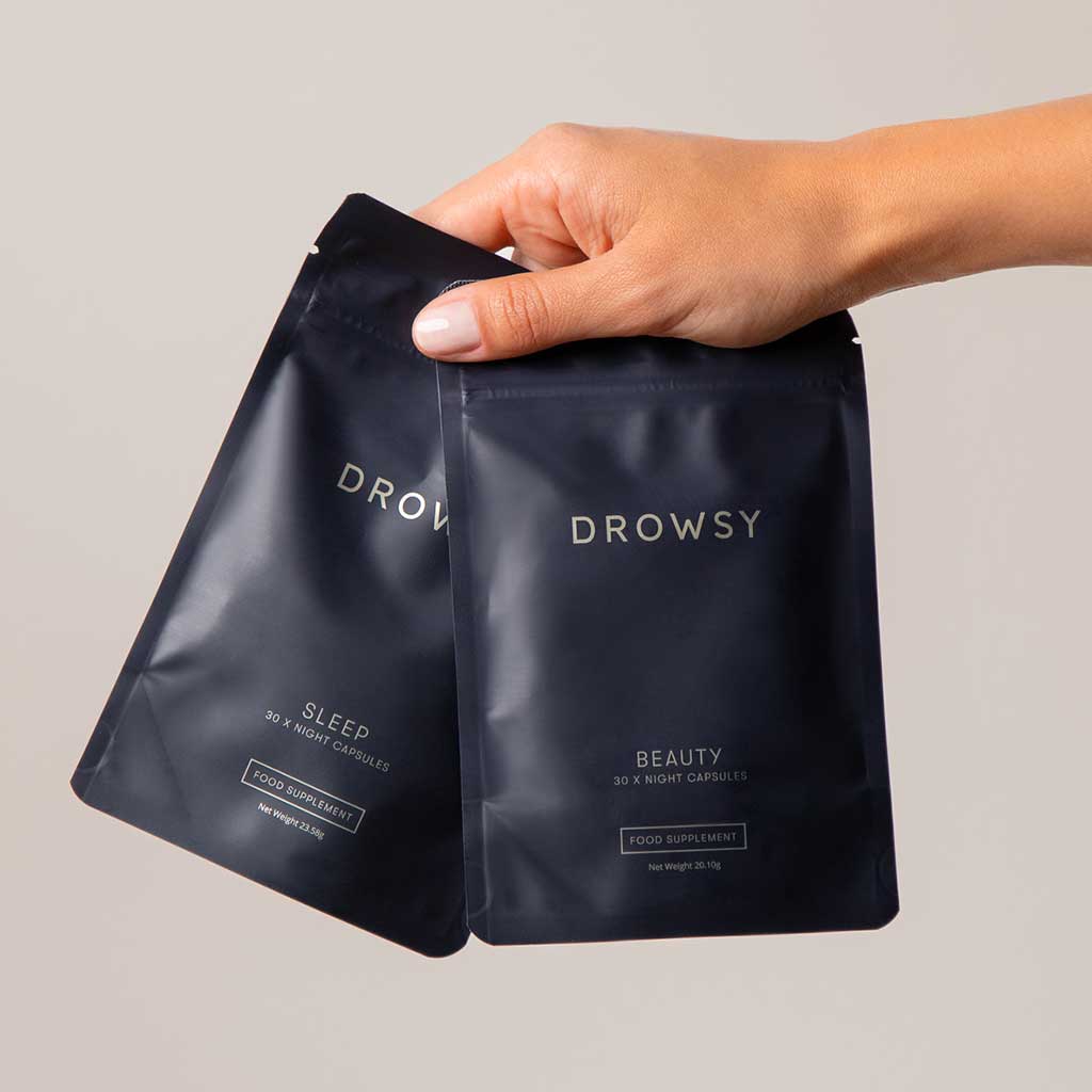 A hand holds two drowsysleepco packets from the Beauty Sleep Clinic Subscription: one labeled Sleep, 30 night capsules and another Beauty, 30 x night capsules. Both are food supplements shown against a plain background.