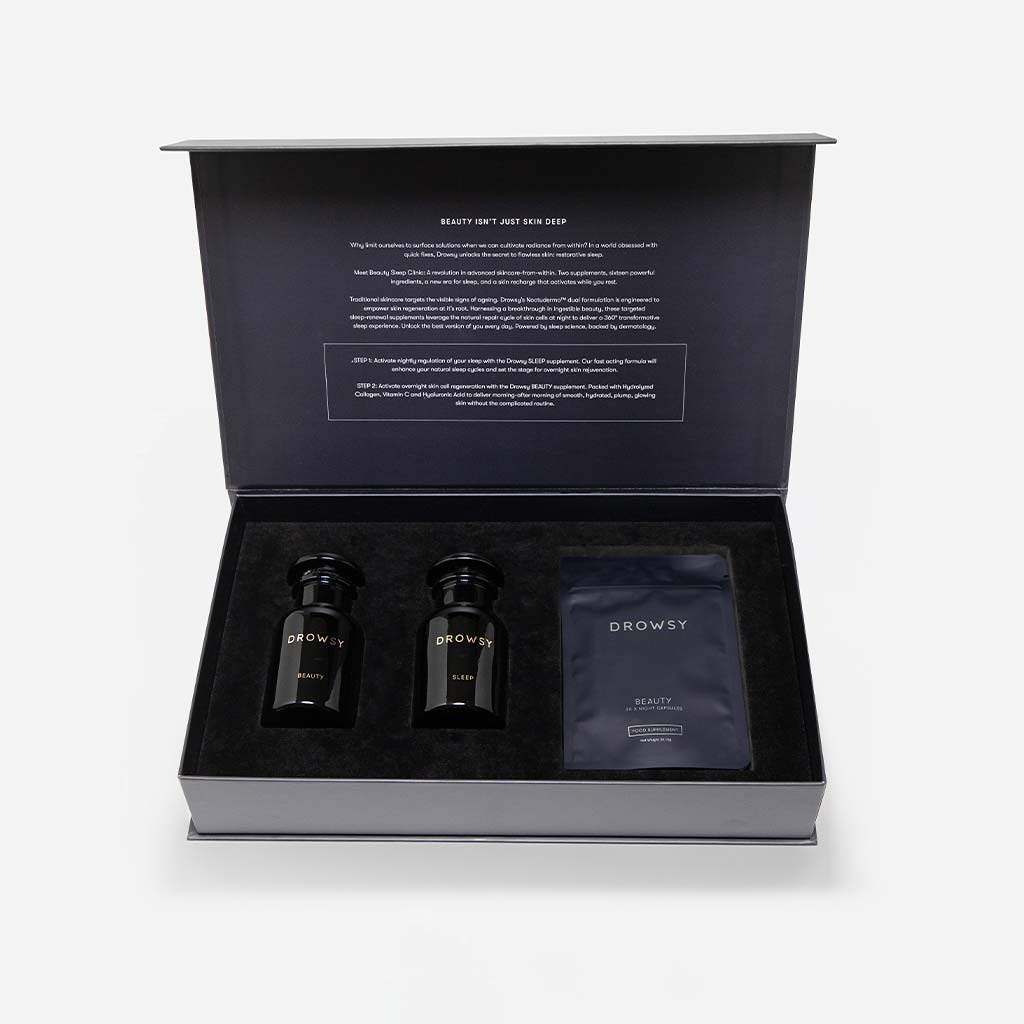 A sleek black gift box from drowsysleepco contains two small black bottles from the Supplements - Sleep & Glow Protocol and a packet labeled Drowsy Beauty, all nestled in black foam with a product description printed inside the lid.
