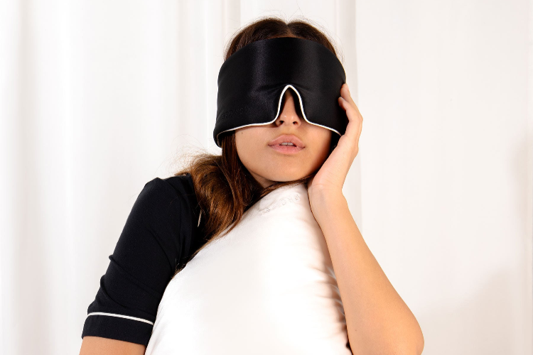 A person wearing a black sleep mask holds a white pillow and gently touches their face, standing against a white background.