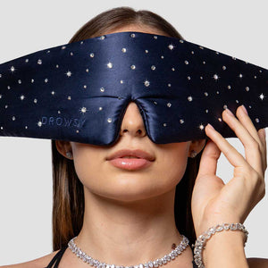 A woman wears the drowsysleepco Silk Sleep Mask Crystals by Swarovski®, covering her eyes and forehead, while holding it with one hand and accessorizing with a sparkly necklace, earrings, and bracelet.