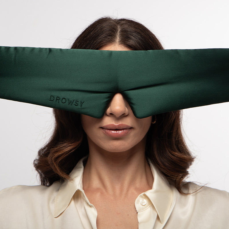 A person wearing a wide, dark green silk Ivy Garden sleep mask from drowsysleepco over their eyes.