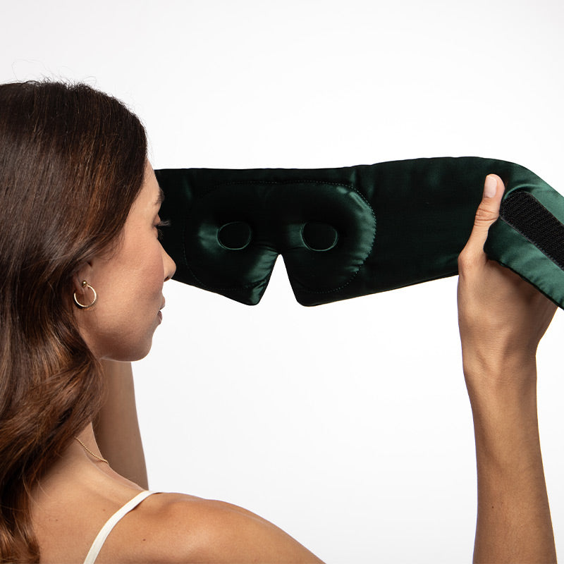 A person holds a dark green Ivy Garden - Eyelash Protecting Mask, a contoured silk sleep mask, in front of their face.