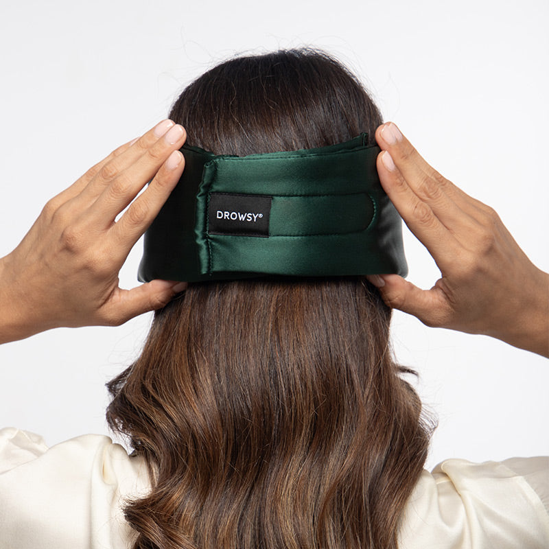 A person with long brown hair adjusting a dark green Ivy Garden silk sleep mask by drowsysleepco, featuring a velcro closure at the back of their head.