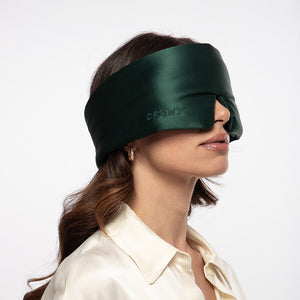 A person wearing a dark green Ivy Garden silk sleep mask over their eyes.