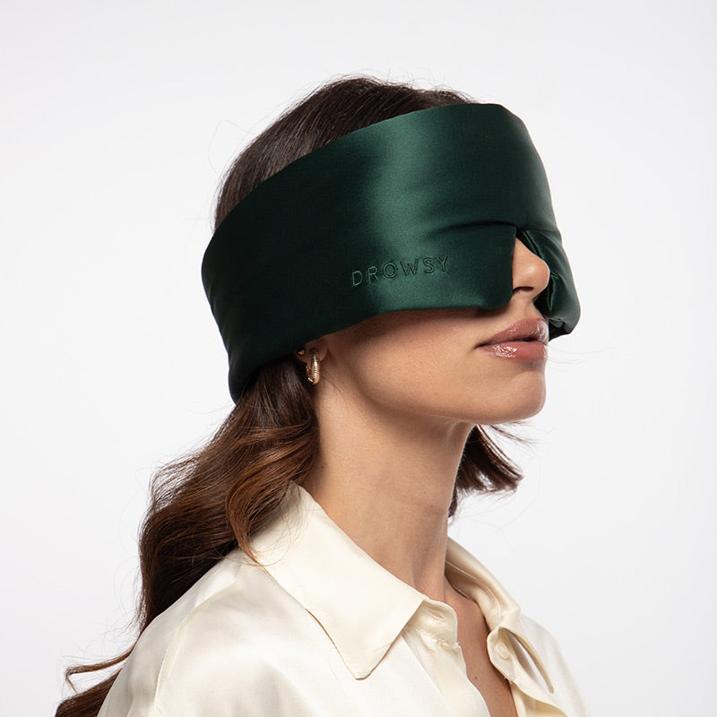 A person wearing a dark green Ivy Garden silk sleep mask over their eyes.
