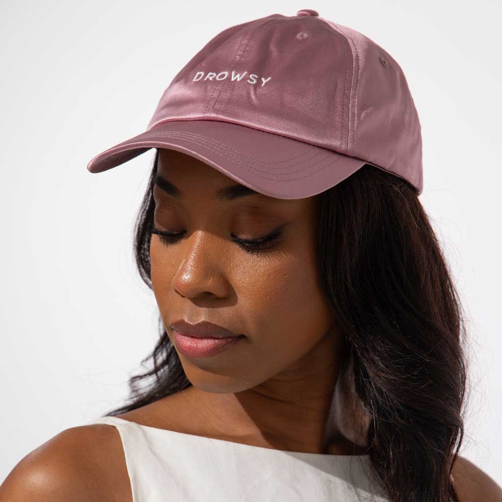 A woman with long dark hair wears the Drowsy Silk Cap in Damask Rose by Drowsy and a white sleeveless top, looking down with a calm expression against a plain light background.