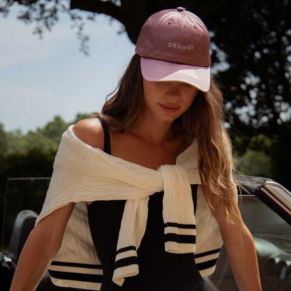 A woman in a midnight blue DROWSY cap from drowsysleepco’s Perfect Escape Bundle stands by a car, draped in a white sweater over a black top, surrounded by lush greenery—embodying beauty sleep style.