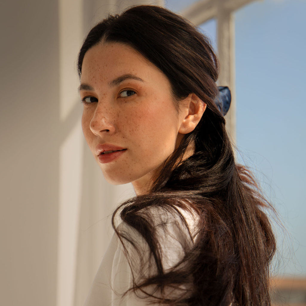 A woman with long dark hair, fair skin, and freckles looks over her shoulder in natural sunlight near a window. She is wearing a white top and has her hair tied back with a blue hair clip.