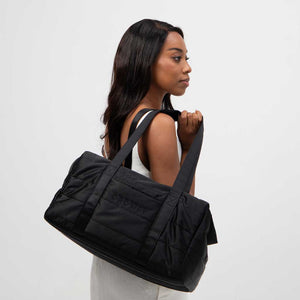 A woman in a white dress stands sideways holding the drowsysleepco Perfect Escape Bundle - Black Jade duffel bag, ideal for carrying your favorite sleep mask, set against a plain white background.