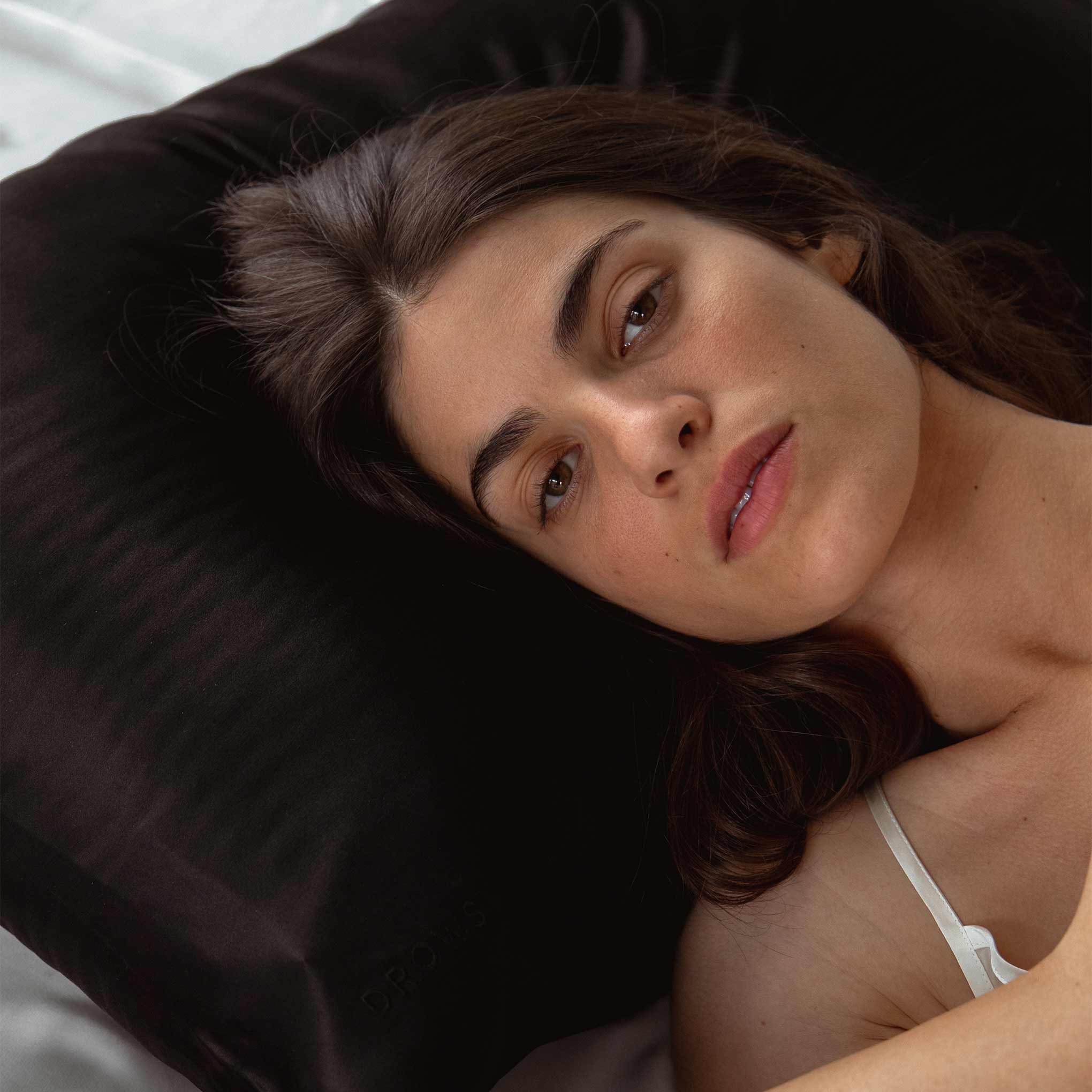 A woman with long dark hair, wearing a white strap top, relaxes on the drowsysleepco Sensual Sleep Bundle - Black Jade pillowcase, gazing at the camera in soft natural light.