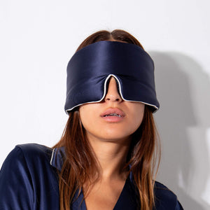 A woman wears the drowsysleepco Midnight Blue - Pipe Dreams Edition silk sleep mask and matching pajamas against a white background. The adjustable mask covers her eyes and part of her nose, with her brown hair visible.